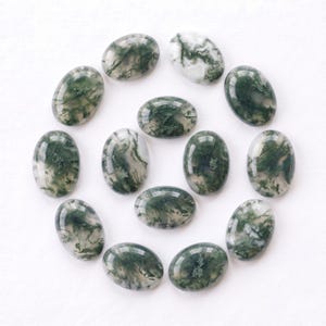 Pu&ograve; includere: Una collezione di tredici cabochon di agata muschiata di forma ovale disposti in uno schema circolare. Ogni pietra lucidata presenta una base bianca traslucida con distinte inclusioni dendritiche verde scuro simili a felci.