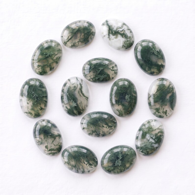 Pu&ograve; includere: Una collezione di tredici cabochon di agata muschiata di forma ovale disposti in uno schema circolare. Ogni pietra lucidata presenta una base bianca traslucida con distinte inclusioni dendritiche verde scuro simili a felci.