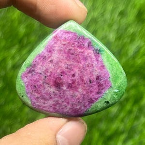 以下が含まれることがあります： 鮮やかな色の宝石。中心部は質感のある濃いピンク色で、明るい緑色の縁で囲まれています。宝石は緑の草を背景に保持されており、その自然な美しさを際立たせています。
