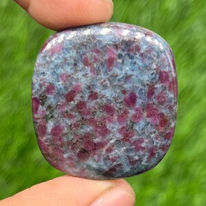 Puede incluir: Una piedra preciosa pulida, de forma cuadrada, con un patrón moteado. La piedra presenta una mezcla de tonos azul claro, gris y rojo intenso. El rojo aparece en pequeñas manchas irregulares, creando una textura visual única.