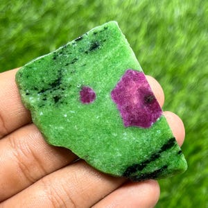 May include: A rough-cut gemstone featuring a vibrant green base with black veining and two prominent ruby inclusions. The ruby inclusions are a deep magenta color, with one appearing as a small circle and the other as a larger, angular shape.