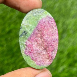 May include: An oval-shaped gemstone with a vibrant color combination. The stone features a striking blend of pink and green hues, with the pink dominating the center and the green forming a border. The stone is held between fingers.