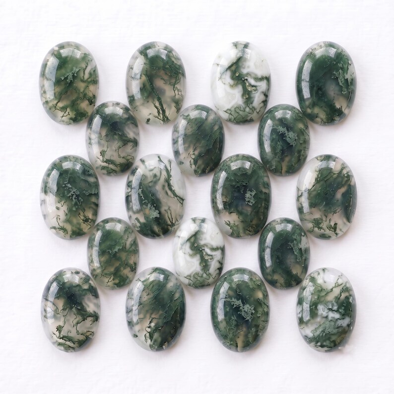 Pu&ograve; includere: Una collezione di quindici cabochon di agata muschiata di forma ovale disposti in una griglia. Ogni pietra lucidata presenta una base bianca traslucida con distinte inclusioni dendritiche verde scuro che creano motivi organici unici.