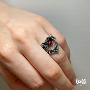 May include: A silver-toned ring featuring a V-shaped band with black accents and a central design. The design includes a large, round, red gemstone and crescent moon details. The ring is worn on a finger.