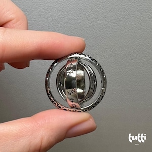 May include: A silver-colored, spherical ring with multiple rotating bands. The outer ring has an ornate, patterned design. The inner bands feature engraved characters. The ring is held between two fingers against a gray background.