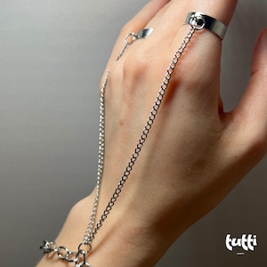 Puede incluir: Una cadena de mano plateada con un anillo y un anillo para el dedo conectados por una delicada cadena. La joya es de metal brillante y está diseñada para usarse en la mano. La cadena también se conecta a una pulsera.
