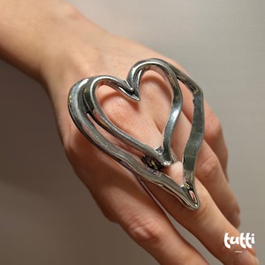 May include: A silver-toned ring in the shape of a heart. The ring is worn on a finger and has a three-dimensional design. The heart shape is open and airy, with smooth, curved lines. The ring is a statement piece.