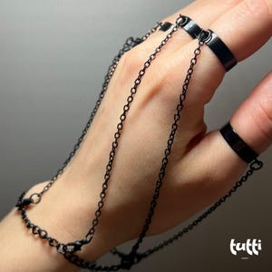 May include: Black hand chain jewelry featuring three rings connected by delicate chains. The rings are worn on the fingers and connected to a bracelet-like chain that drapes over the back of the hand. The jewelry is made of a dark metal.