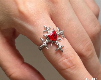 Peut inclure: Une bague argentée ornée d'un cœur rouge en son centre. La bague présente un motif en forme de croix avec de petits détails pointus s'étendant vers l'extérieur. La bague est portée au doigt.