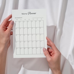 Può includere: Un'agenda di carta bianca intitolata "Mama Planner" con un layout a griglia per la pianificazione mensile. L'agenda è tenuta in mano su uno sfondo di tessuto bianco. Il testo "Mama Planner" è in un carattere corsivo.