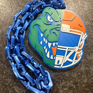 May include: A blue chain is attached to a two-part emblem. One half shows a green alligator head with white teeth, and the other half is an orange and blue American football helmet. The emblem is a sports-themed design.