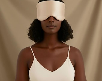 May include: A woman wearing a light beige sleep mask. The mask covers the eyes and is made of a smooth, satin-like material. She is wearing a matching camisole. The background is a neutral beige color.