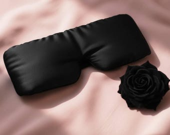 May include: A black sleep mask made of a soft, silky material. A black rose is next to the mask. The mask is designed to block out light for better sleep. The items are on a pink satin surface.