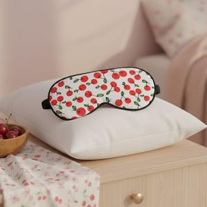 Peut inclure: Un masque de sommeil blanc orné de motifs de cerises rouges et de feuilles vertes, posé sur un oreiller blanc. Le masque a un bord noir et des sangles élastiques. L'image évoque une ambiance de chambre à coucher.