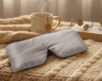 May include: A gray satin eye mask rests on a beige cable-knit blanket. A cup of tea with steam rising from it and a book are in the background. The eye mask is designed to block out light.