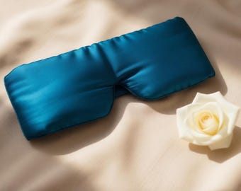 May include: A teal-colored satin sleep mask with a curved design, resting on a beige fabric surface. A white rose with a yellow center is placed near the mask. The mask is designed to block out light for better sleep.