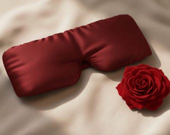 May include: A deep red sleep mask made of a silky material, lying on a cream-colored fabric. A red rose is placed next to the mask. The sleep mask is designed to block out light for better sleep.