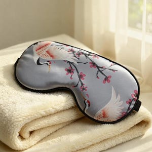 May include: A gray satin sleep mask with a black trim, featuring a crane and floral design in pink and white. The mask rests on a folded, cream-colored blanket.