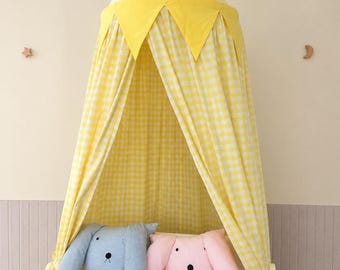 Può includere: Un baldacchino in tessuto a quadretti gialli e bianchi con una parte superiore a punta e gagliardetti triangolari gialli appesi sopra una culla bianca. Due lunghi cuscini in peluche a forma di coniglio, uno azzurro e uno rosa, sono nella culla.