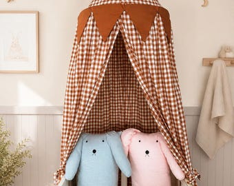 Può includere: Un baldacchino in tessuto a quadretti marrone e bianco con una parte superiore a punta e gagliardetti triangolari marrone tinta unita appesi al bordo. Due lunghi cuscini in peluche, uno azzurro e uno rosa chiaro, hanno occhi e nasi ricamati.