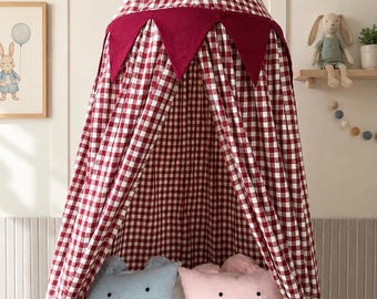 Può includere: Un baldacchino conico con motivo a quadretti vichy rossi e bianchi, rifinito con un bordo di bandierine triangolari rosso scuro lungo la parte superiore. Il baldacchino pende sopra una culla bianca contenente due cuscini rettangolari, azzurro e rosa.