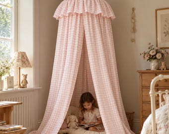 Può includere: Un baldacchino in tessuto sospeso al soffitto con un motivo a quadretti vichy rosa chiaro e bianco. Il baldacchino presenta una parte superiore conica con un bordo arricciato e lunghi pannelli fluidi che scendono fino a terra, creando uno spazio a tenda.