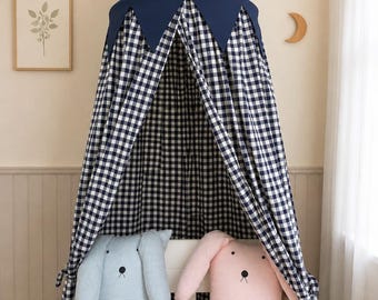 Può includere: Un baldacchino in tessuto a quadretti blu navy e bianco pende sopra una culla bianca. Due cuscini di peluche dalle orecchie lunghe, uno azzurro e uno rosa pallido, ciascuno caratterizzato da un semplice viso ricamato con occhi e naso neri.