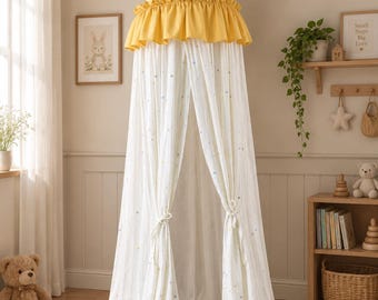Può includere: Un baldacchino in tessuto che arriva fino a terra, caratterizzato da una parte superiore conica a righe giallo senape e bianche con un bordo arricciato. Il corpo principale è in tessuto bianco decorato con piccole stelle multicolori e due lacci.
