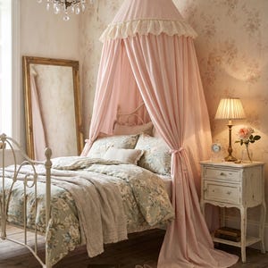 K&ouml;nnte beinhalten: Ein hellrosa Betthimmel aus Stoff mit ger&uuml;schtem Rand h&auml;ngt &uuml;ber einem wei&szlig;en Metallbettgestell. Das Bett ist mit einer floral gemusterten Bettw&auml;sche auf hellgr&uuml;nem Grund bezogen. Ein passender wei&szlig;er Nachttisch und ein hoher Spiegel sind zu sehen.