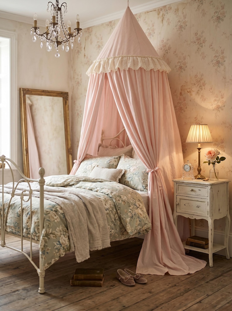 K&ouml;nnte beinhalten: Ein hellrosa Betthimmel aus Stoff mit ger&uuml;schtem Rand h&auml;ngt &uuml;ber einem wei&szlig;en Metallbettgestell. Das Bett ist mit einer floral gemusterten Bettw&auml;sche auf hellgr&uuml;nem Grund bezogen. Ein passender wei&szlig;er Nachttisch und ein hoher Spiegel sind zu sehen.