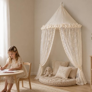 May include: A cream-coloured fabric canopy with a conical top and lace curtains featuring pom-pom trim. The canopy hangs over a round, tufted floor cushion accompanied by two square throw pillows and a small plush rabbit toy on a light wood floor.