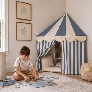 Könnte beinhalten: Ein Eckspielzelt mit einem blau-weiß gestreiften Stoffdach und einer gewellten Kante. Das Zelt verfügt über ein spitzes Dach und Vorhänge zum Zusammenbinden, die einen kleinen Innenraum mit sichtbaren Regalen und einem Bodenkissen bilden.