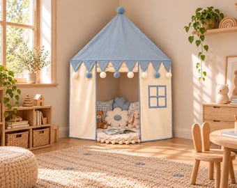 Può includere: Una tenda da gioco in tessuto color crema e azzurro con tetto a punta sormontato da un pompon, mantovane triangolari con pompon alternati blu e bianchi e una finestra quadrata. L'interno contiene cuscini e animali di peluche.