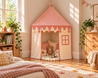 Può includere: Una tenda da gioco a quadretti rosa e bianchi con tetto a punta, bordo con pon pon e una finestra quadrata. L'interno presenta cuscini morbidi e peluche, tra cui un orsetto e un coniglio, disposti su un tappeto rotondo con pon pon.