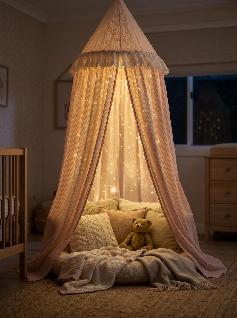 K&ouml;nnte beinhalten: Ein hellrosa Stoffhimmel h&auml;ngt von der Decke, mit einem ger&uuml;schten oberen Rand und innenliegenden Lichterketten. Im Inneren befindet sich ein rundes Bodenkissen, das mit mehreren strukturierten Kissen und einer weichen Decke sowie einem kleinen Teddyb&auml;ren ausgelegt ist.