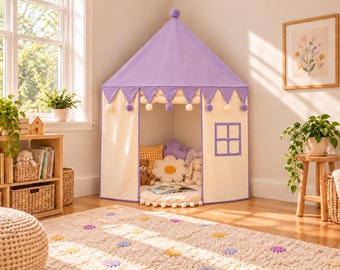 Può includere: Una tenda da gioco in tessuto color viola e crema con tetto a punta, caratterizzata da un bordo smerlato con pompon bianchi, una finestra quadrata e un ingresso aperto. All'interno, la tenda contiene cuscini e peluche.