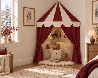 Può includere: Una tenda da gioco angolare con tetto conico a pannelli in tessuto rosso scuro e crema. La tenda presenta mantovane smerlate e tende rosse abbinate, legate lateralmente per rivelare un interno accogliente con cuscini e un orsacchiotto.
