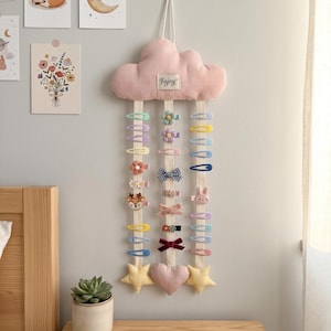 Op de afbeelding: Een roze, wolk-vormige haarcliphouder met het woord "Fijoy" erop, hangend aan een muur. De houder heeft meerdere rijen kleurrijke haarclips, waaronder bloemen, strikken en dierontwerpen. Decoratieve wandkunst is zichtbaar op de achtergrond.