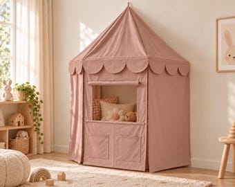 Può includere: Una tenda da gioco in tessuto rosa polvere con tetto a punta dai bordi smerlati, una finestra avvolgibile e due tasche frontali inferiori. All'interno della tenda, due cuscini quadrati e un piccolo orsacchiotto sono visibili.