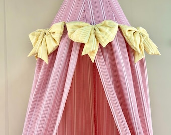 Può includere: Un baldacchino in tessuto di forma conica con un motivo a righe rosse e bianche. La struttura è decorata con tre grandi fiocchi giallo pallido in tessuto a quadretti, posizionati orizzontalmente sulla parte anteriore della tenda sospesa.