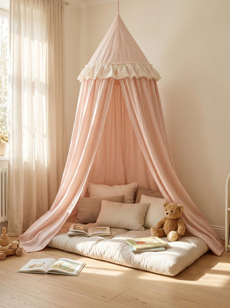May include: A pale pink fabric canopy suspended from the ceiling, featuring a ruffled white eyelet trim at the top. The canopy drapes over a rectangular floor mattress adorned with several neutral-toned pillows, an open book, and a teddy bear.