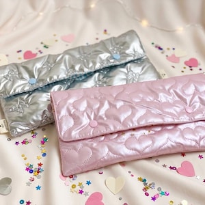 Op de afbeelding: Twee gewatteerde clutches. Eén is zilverkleurig met een sterrenpatroon, de andere roze met een hartjespatroon. Beide hebben een knoopsluiting en een klein label. De tassen liggen op een licht oppervlak met confetti en hartvormige decoraties.