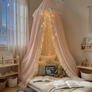 K&ouml;nnte beinhalten: Ein hellrosa Betthimmel aus Stoff mit wei&szlig;em R&uuml;schenbesatz h&auml;ngt von der Decke und &uuml;berspannt eine Bodenmatratze mit Kissen, einem Teddyb&auml;ren und offenen B&uuml;chern. Eine Lichterkette mit warmwei&szlig;em Licht ist in den Stoff des Himmels eingearbeitet.