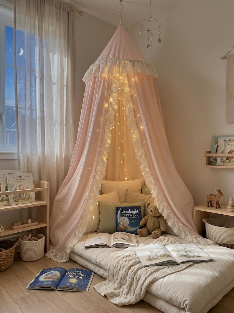 K&ouml;nnte beinhalten: Ein hellrosa Betthimmel aus Stoff mit wei&szlig;em R&uuml;schenbesatz h&auml;ngt von der Decke und &uuml;berspannt eine Bodenmatratze mit Kissen, einem Teddyb&auml;ren und offenen B&uuml;chern. Eine Lichterkette mit warmwei&szlig;em Licht ist in den Stoff des Himmels eingearbeitet.