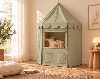 Può includere: Una tenda da gioco in tessuto verde salvia con tetto a punta e bordo smerlato, una finestra rettangolare e due tasche inferiori. All'interno si trovano un cuscino a quadri marroni, un cuscino floreale e un coniglietto di peluche.