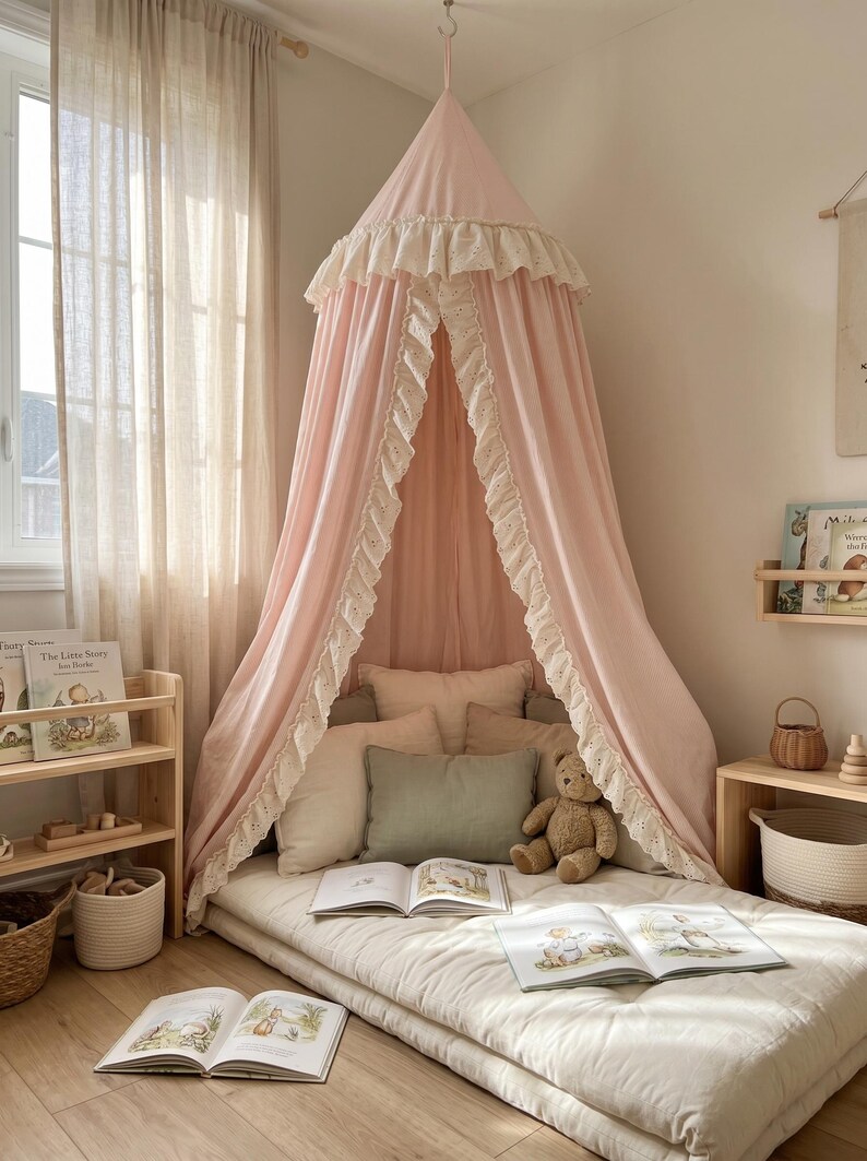 K&ouml;nnte beinhalten: Ein hellrosa Stoffhimmel mit wei&szlig;er &Ouml;senstickerei an den R&auml;ndern, der von der Decke h&auml;ngt. Der Betthimmel &uuml;berspannt eine Bodenmatratze mit mehreren Kissen, einem braunen Teddyb&auml;ren und aufgeschlagenen Bilderb&uuml;chern auf der Liegefl&auml;che.