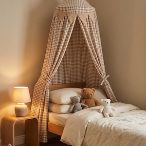May include: A beige and white gingham fabric bed canopy featuring a conical top and a decorative row of triangular pennants. The canopy hangs over a wooden bed frame with white bedding, three plush teddy bears, and a bedside lamp on a wooden table.