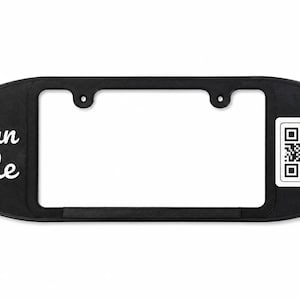 May include: A black, oval-shaped license plate frame featuring the white text "Scan Me" on the left side and a square black and white QR code on the right side. The frame has two mounting holes at the top for attachment to a vehicle.