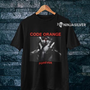 Könnte beinhalten: Schwarzes T-Shirt mit dem roten Schriftzug "CODE ORANGE" über einem Schwarz-Weiß-Bild. Darunter steht das Wort "FOREVER" in Rot. Das T-Shirt hängt an einem Holzkleiderbügel.