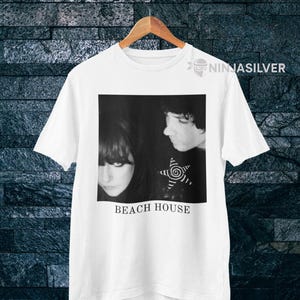 May include: White t-shirt featuring a black and white image of two people with the text "BEACH HOUSE" below. The image includes a star design. The shirt is hanging on a wooden hanger.